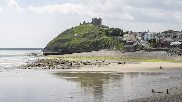 Un pueblo costero con una colina destacada que presenta ruinas de un castillo, una playa arenosa y una hilera de edificios a lo largo de la costa.