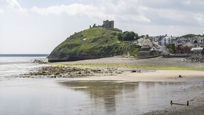 Eine Küstenstadt mit einem markanten Hügel, auf dem eine Burgruine steht, einem Sandstrand und einer Reihe von Gebäuden entlang der Küste.