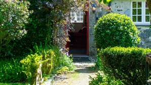 Ein steiniger Pfad führt zu einem Cottage mit roter Tür, umgeben von üppigem Grün und einem gepflegten Garten.