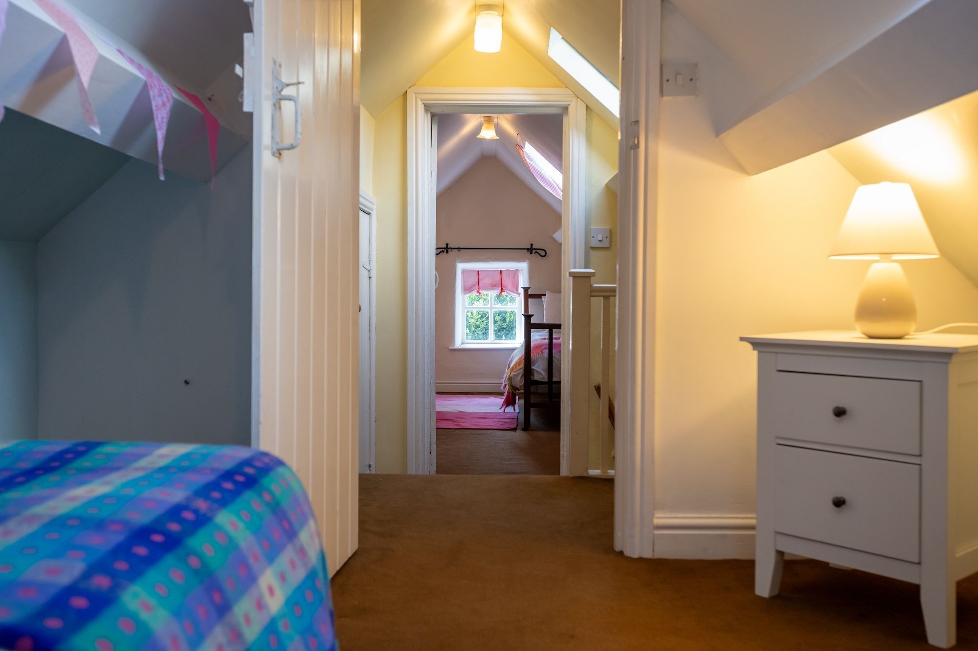 Un dormitorio con cama, mesita de noche, ventana con cortinas y puerta que da a otra habitación.