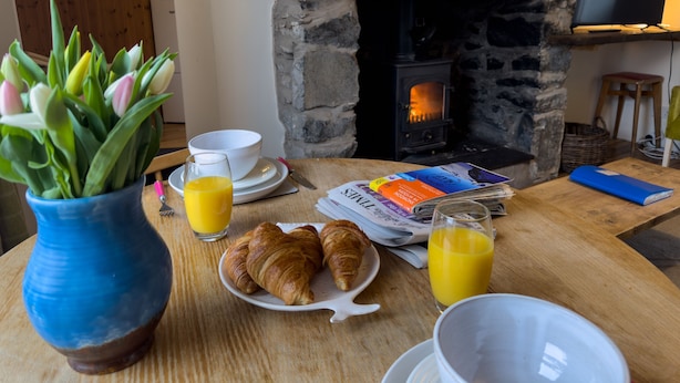 Un desayuno con croissants, jugo de naranja y un periódico sobre una mesa de madera, con una chimenea de piedra al fondo.