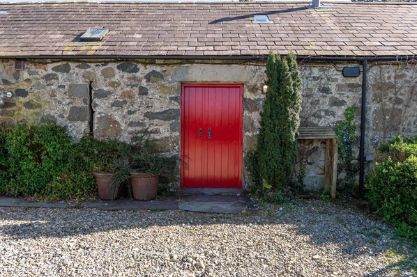 Un bâtiment en pierre avec une porte rouge, des plantes en pot et une allée gravillonnée.