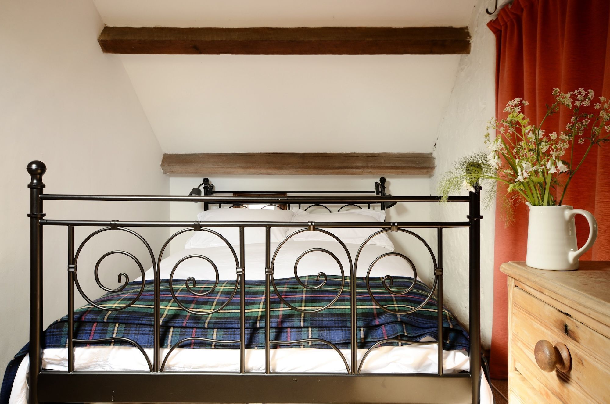 Una cama con estructura metálica y una manta escocesa, un aparador de madera y un florero con flores.