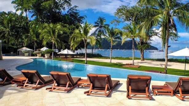 A poolside area with lounge chairs, palm trees, and a view of the ocean. - Recently Renovated • Pacifico L604 Casa Xanadu • 2 BR Ground Floor Condo • Playas del Coco (Playas del Coco)