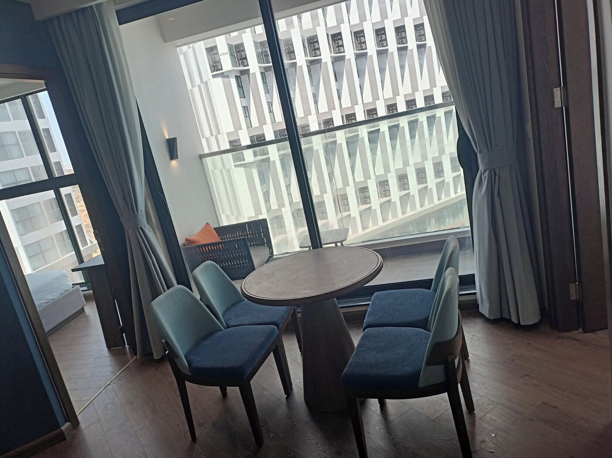 A modern room with a round table and four chairs, large windows with curtains, and a view of a building with balconies.