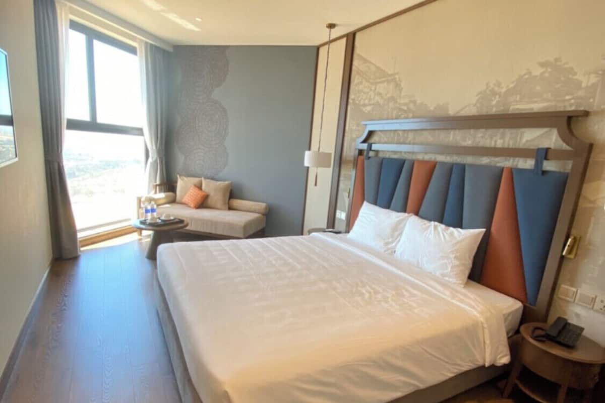 A modern hotel room with a large bed, a seating area with a sofa and armchairs, a small table, and a view of the outdoors.