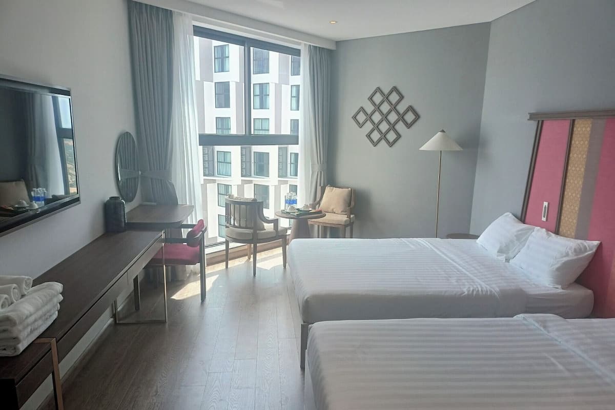 A hotel room with two beds, a desk, a chair, a lamp, and a large window.
