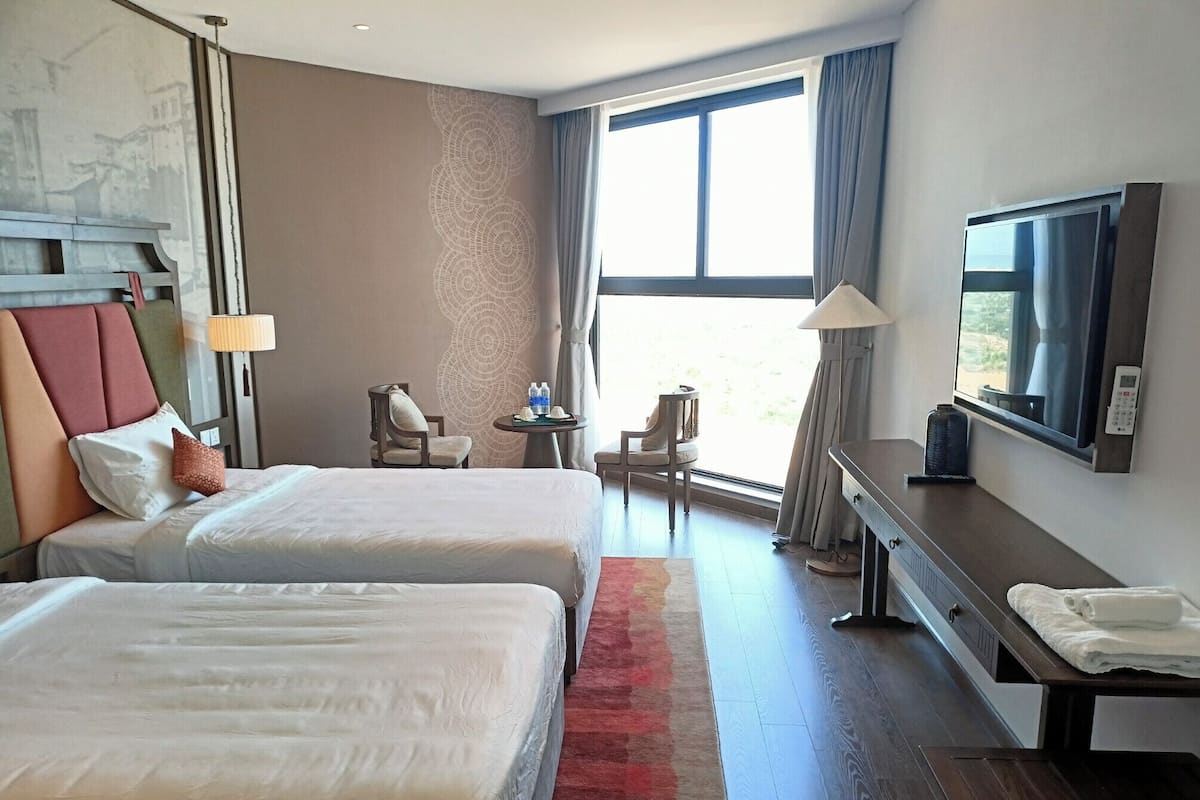 A hotel room with two beds, a desk, a chair, a TV, and a window with curtains.