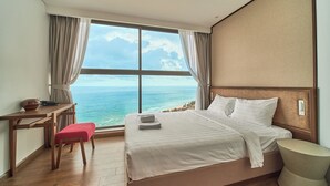 Une chambre d’hôtel dotée d’une grande fenêtre donnant sur la mer, d’un lit avec du linge de maison blanc, d’un bureau en bois avec un bol et d’un petit tabouret.