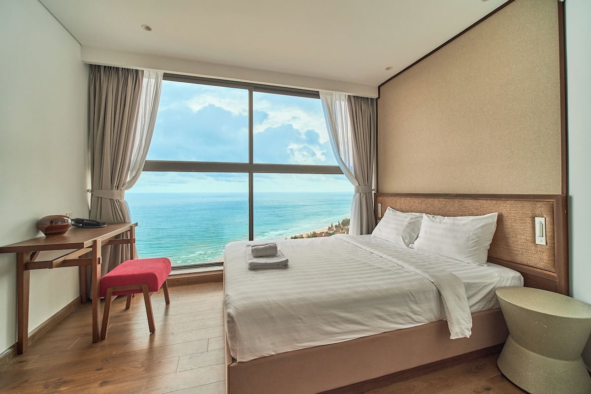 A hotel room with a large window offering a view of the sea, a bed with white linens, a wooden desk with a bowl, and a small stool.