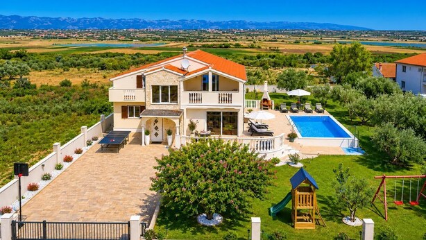 Una casa de dos pisos con piscina, cancha de tenis y vista a montañas distantes.