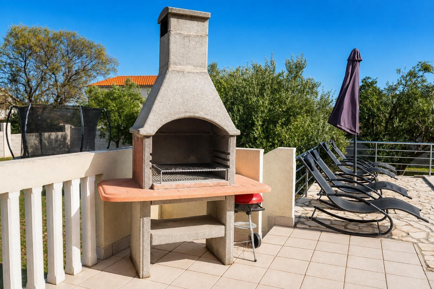 An outdoor stone-built pizza oven with a bench, surrounded by a tiled patio and lounge chairs.
