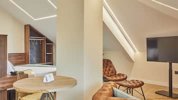 A modern hotel room with a bed, a dining table, a TV, and a seating area with leather chairs.