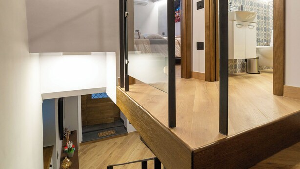 Een moderne trap met houten treden en metalen leuningen, die leidt naar een ruimte op de tweede verdieping met een gewelfd plafond van steen en uitzicht op een slaapkamer.