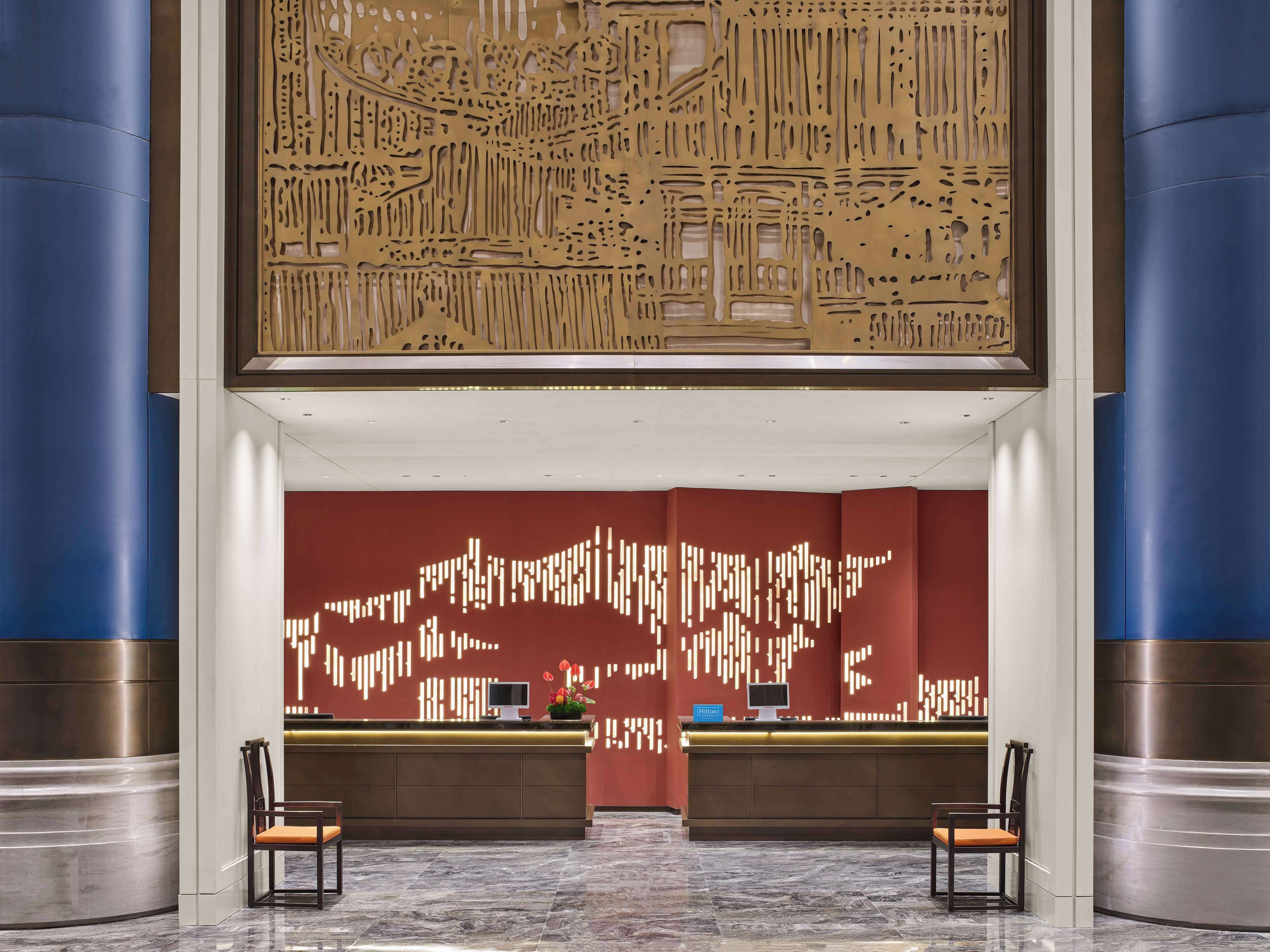 A hotel reception area with a modern design, featuring a reception desk, two chairs, and a decorative wall with abstract patterns.
