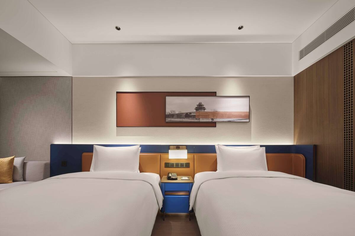Une chambre d’hôtel avec deux lits, une table de chevet avec une lampe, un tableau au mur et un panneau en bois sur le côté droit.