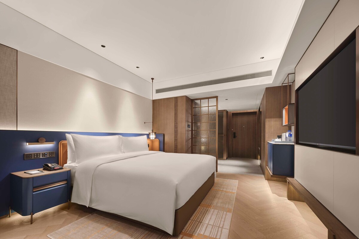 A modern hotel room with a large bed, a bedside table with a phone, a desk with a computer, and a TV on the wall.