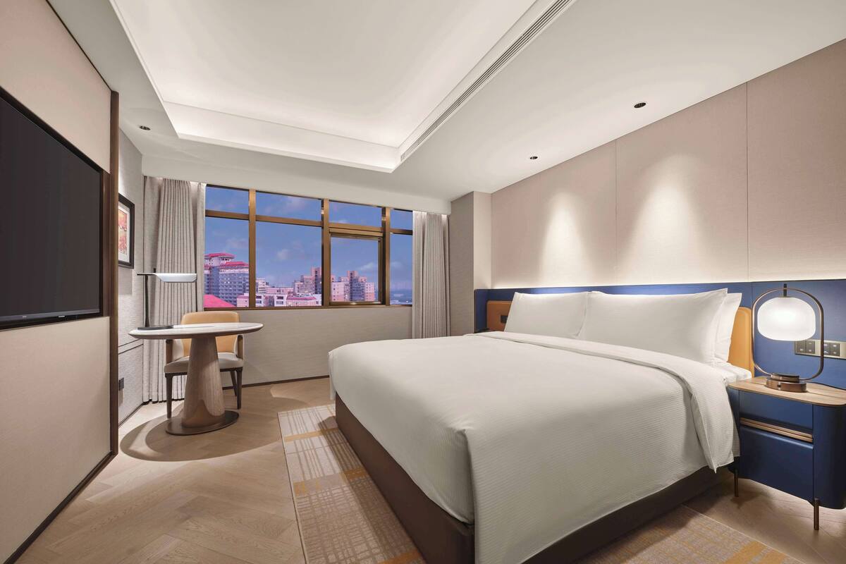 Une chambre d’hôtel avec un grand lit, un bureau, une chaise, une table de chevet et une grande fenêtre donnant sur la ville.