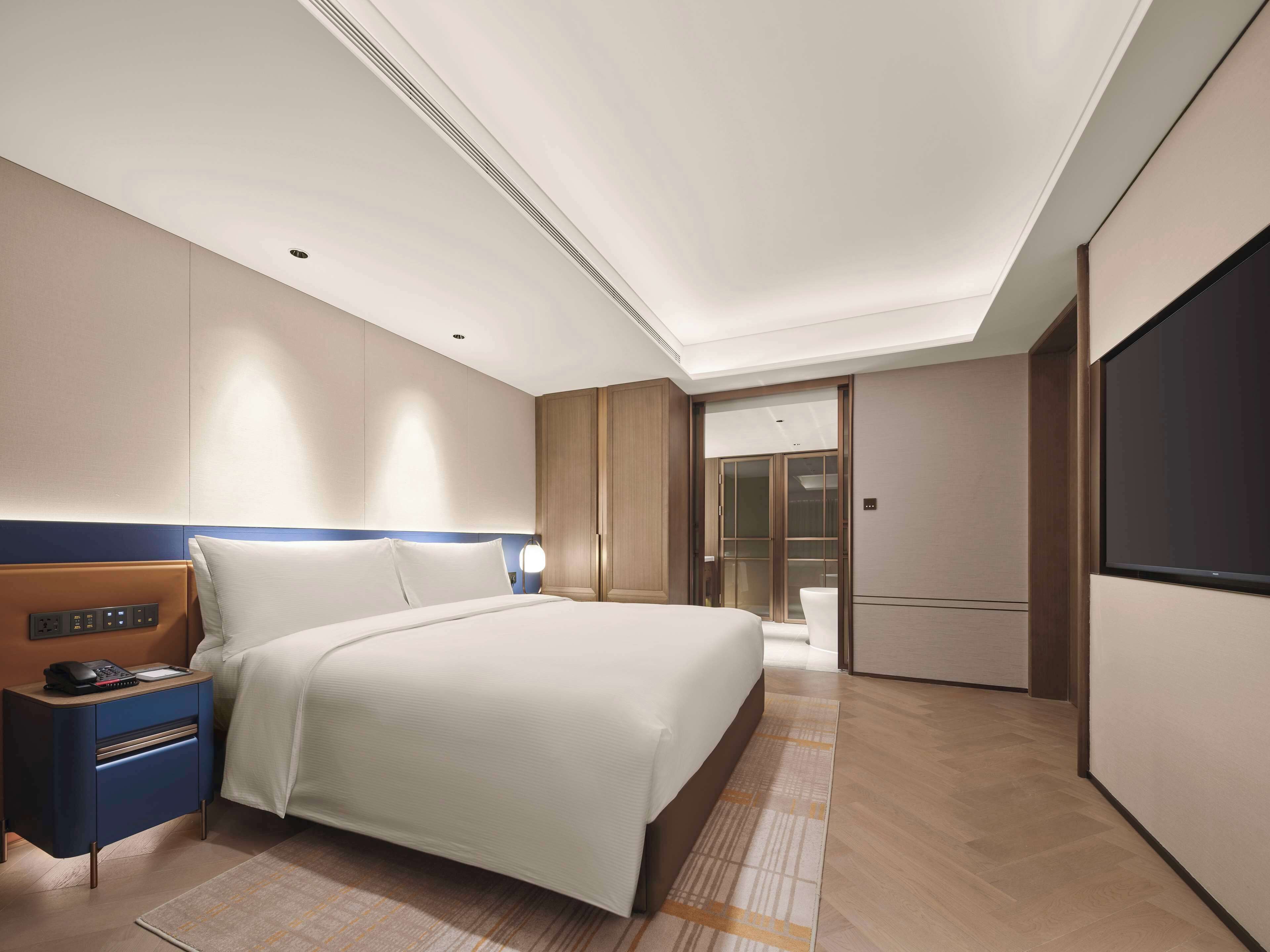 A modern hotel room with a large bed, a bedside table with a phone and charger, a desk with a computer, and a wardrobe.
