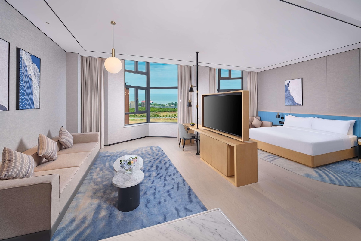 Ein modernes Hotelzimmer mit einem großen Bett, einer Couch, einem Fernseher und Blick ins Freie.