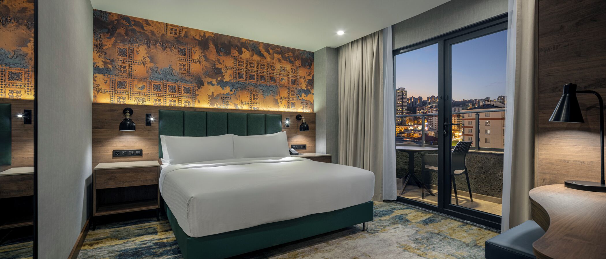 A hotel room with a large bed, a desk, a chair, a view of the city, and a large wall with a patterned design.