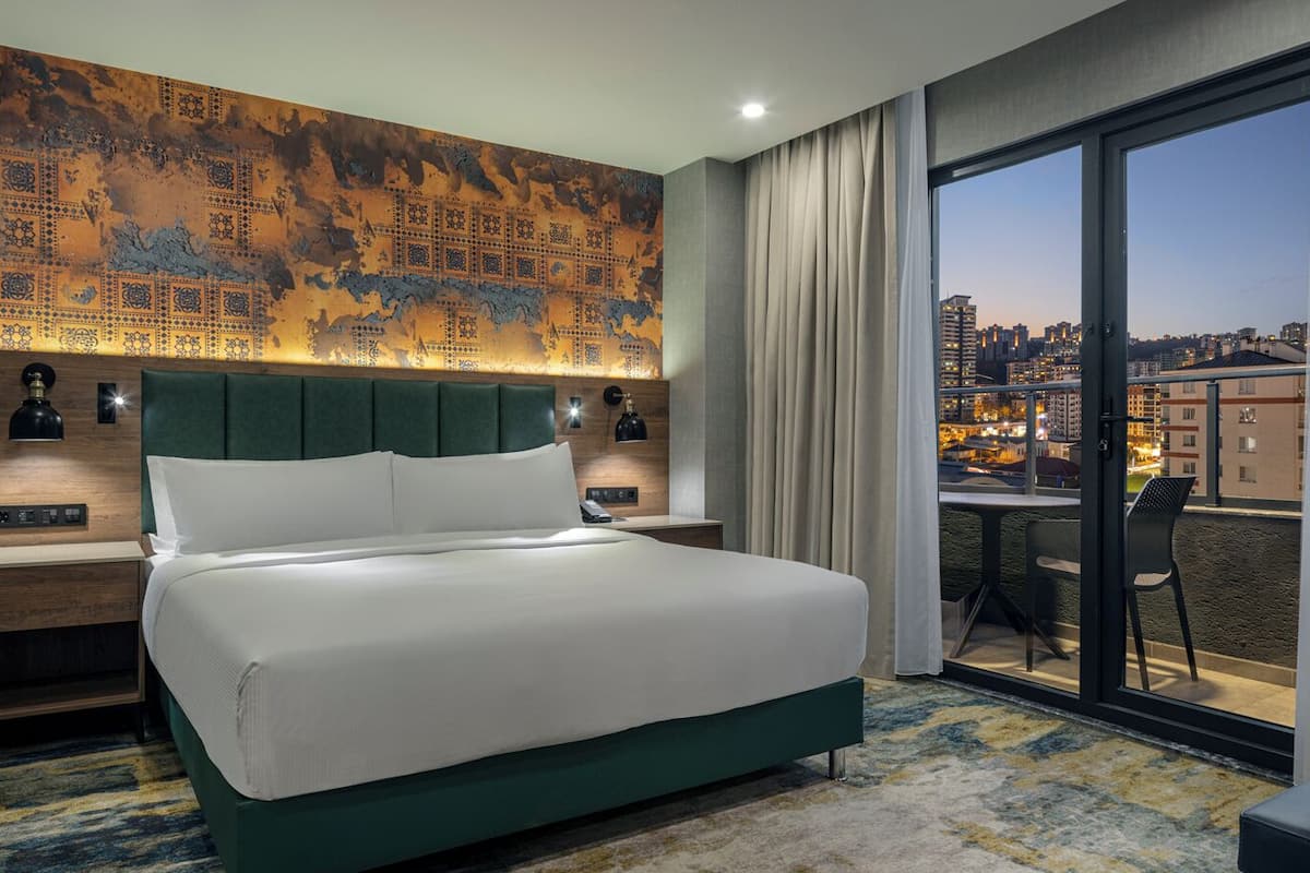 A hotel room with a large bed, a desk, a chair, a view of the city, and a large wall with a patterned design.