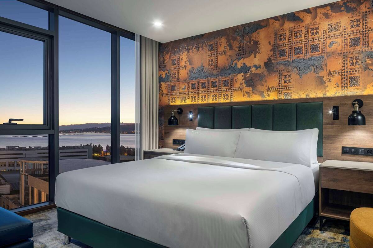 A modern hotel room with a large bed, a view of the cityscape, a patterned wall, and a yellow armchair.