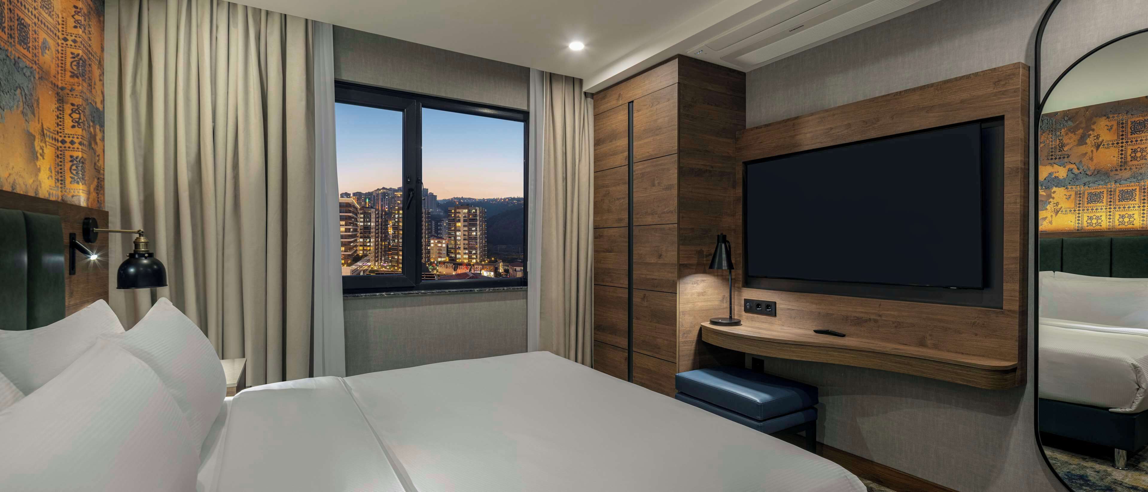 A hotel room with a large bed, a TV mounted on the wall, a desk with a chair, and a window offering a city view.