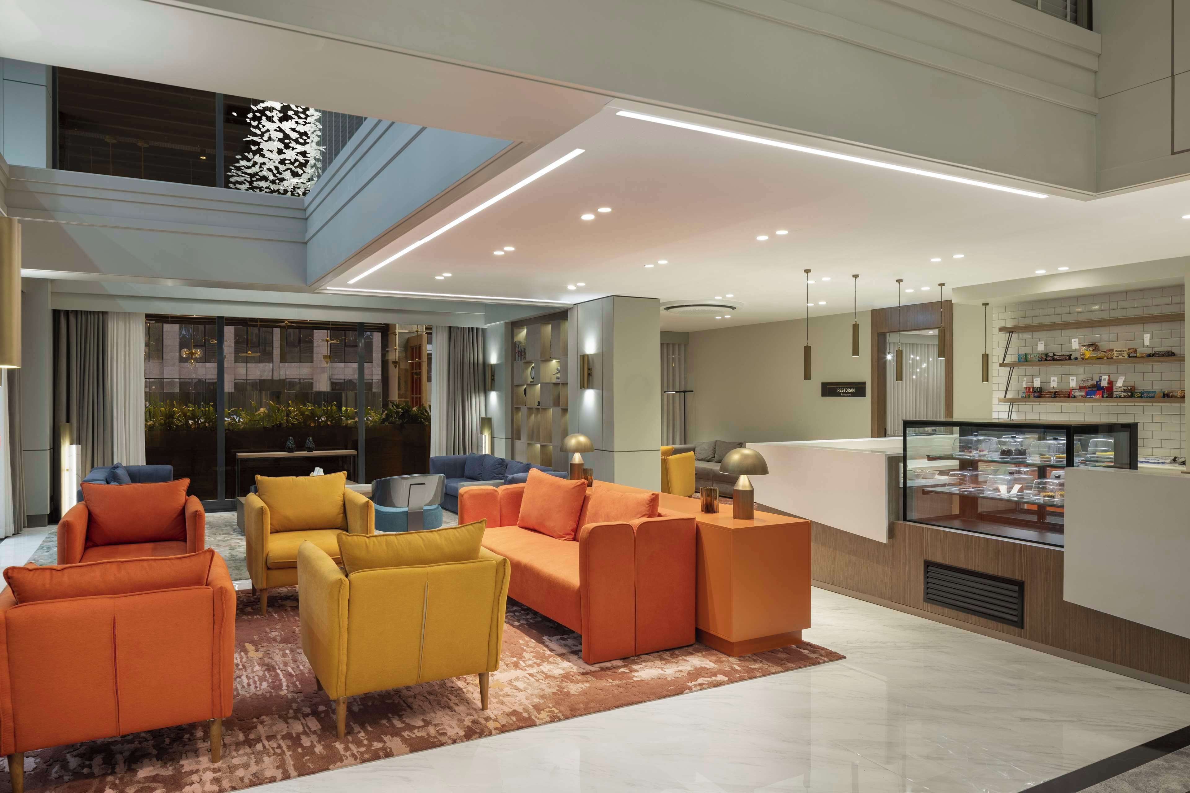 A modern hotel lobby with a seating area featuring orange and yellow sofas, a glass coffee bar, and a view of the cityscape.