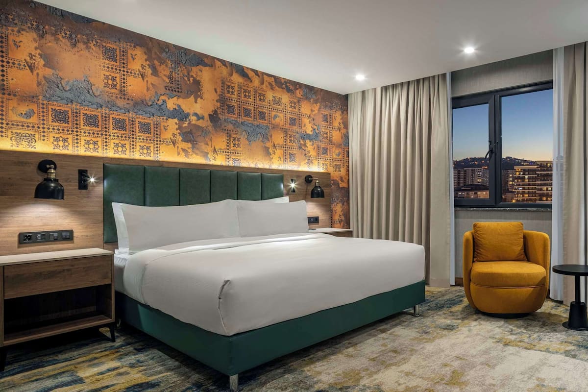 A hotel room with a large bed, a small armchair, a desk, a nightstand, a lamp, and a view of the city at dusk.