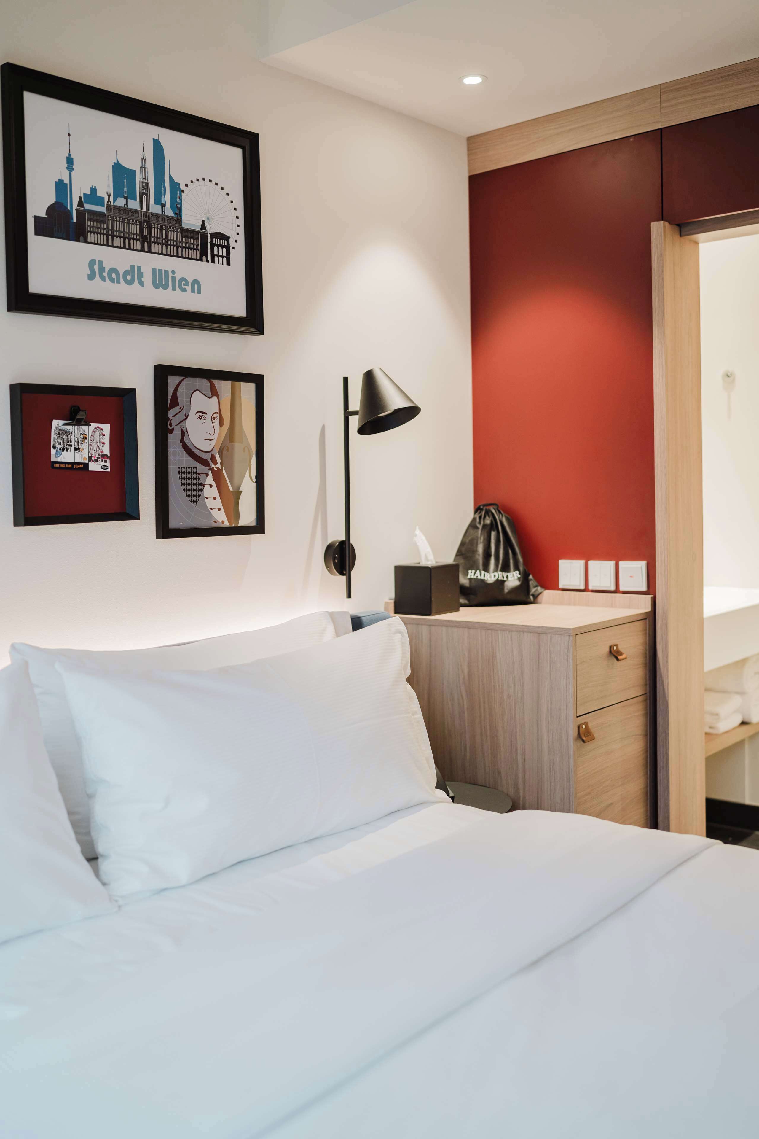 Quarto de hotel com uma cama, uma escrivaninha com luminária, uma parede de destaque vermelha e quadros na parede.