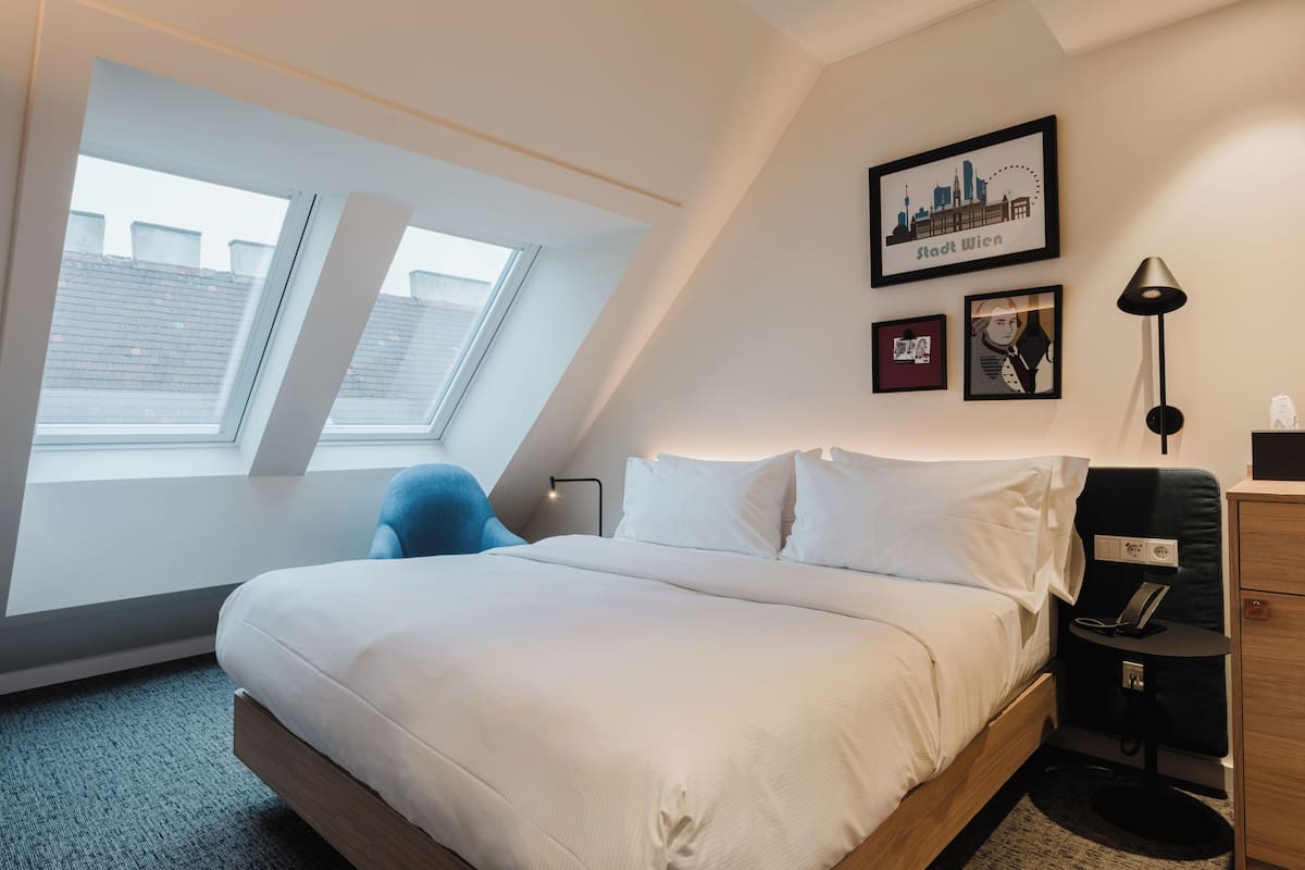 Une chambre d’hôtel moderne avec un grand lit, une lucarne, un bureau avec une lampe et des tableaux encadrés au mur.