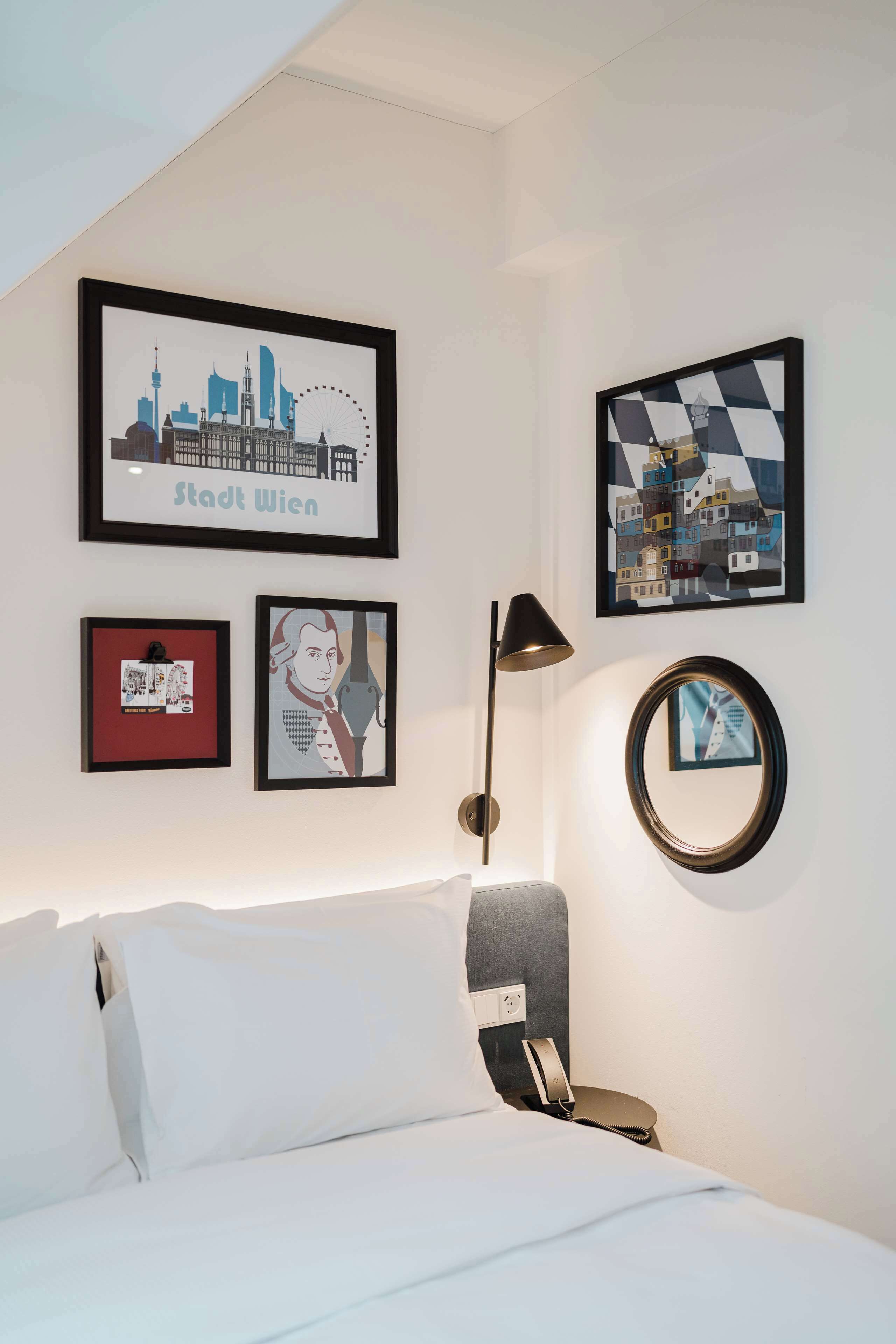 A hotel room with a bed, a wall-mounted lamp, and framed artwork including a cityscape and a portrait.