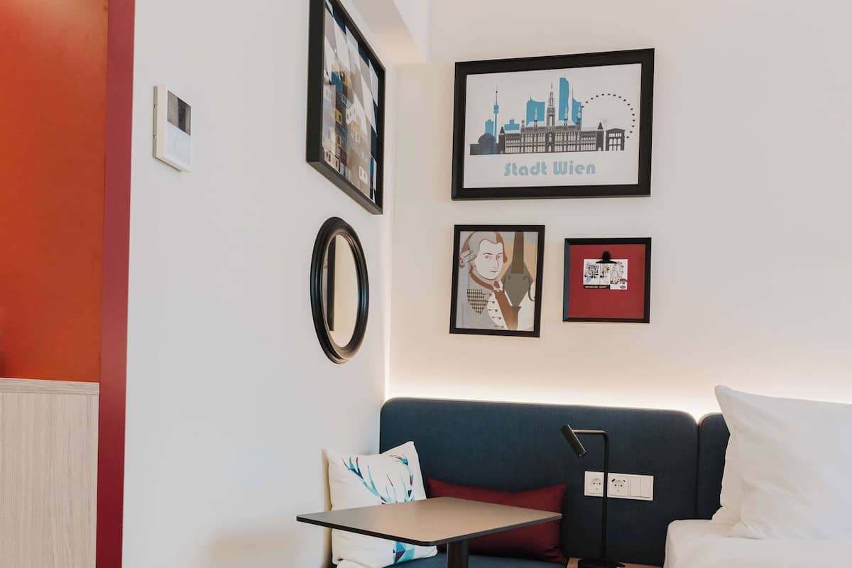 Une chambre d’hôtel avec un lit, un bureau, une chaise et une petite table. Les murs sont ornés de tableaux encadrés et le sol est recouvert d’une moquette.