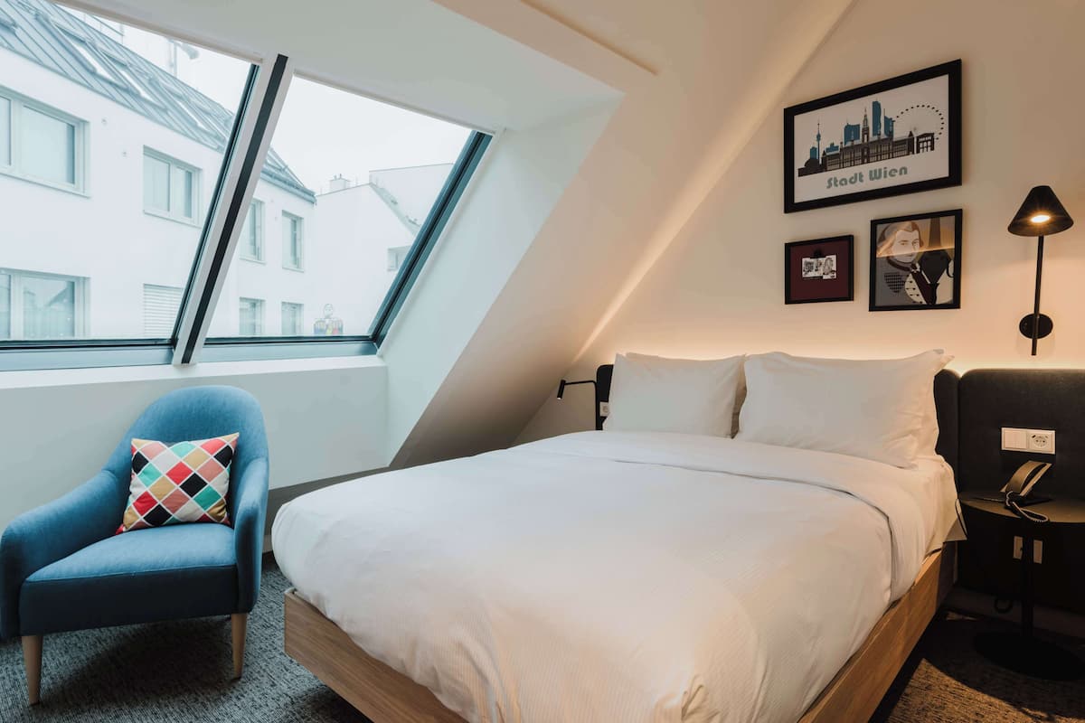 Une chambre d’hôtel moderne avec un grand lit, un fauteuil bleu et un tableau représentant un paysage urbain accroché au mur.