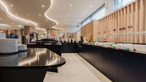 En moderne hotellobby med en buet receptionsdisk, et buffetområde med forskellige madstationer, og en væg beklædt med træpaneler.