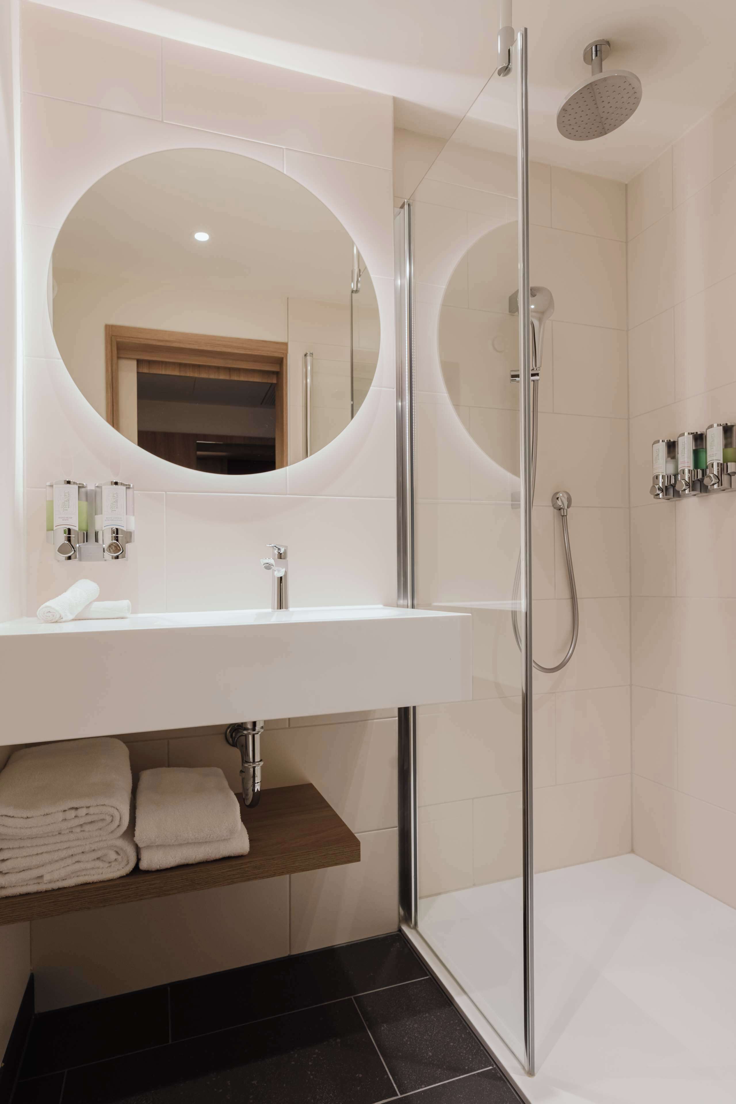 A modern bathroom with a white sink, a round mirror, a glass-enclosed shower, and a towel rack.