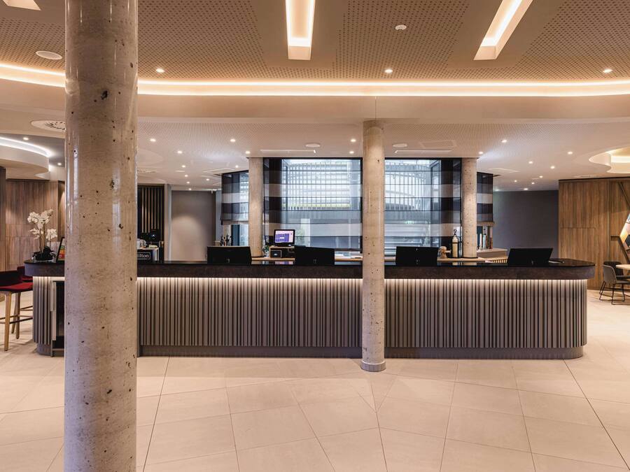 Ein modernes Hotel-Foyer mit einer zentralen Rezeption, Sitzgelegenheiten und einem großen Fenster mit hellem Tageslicht.