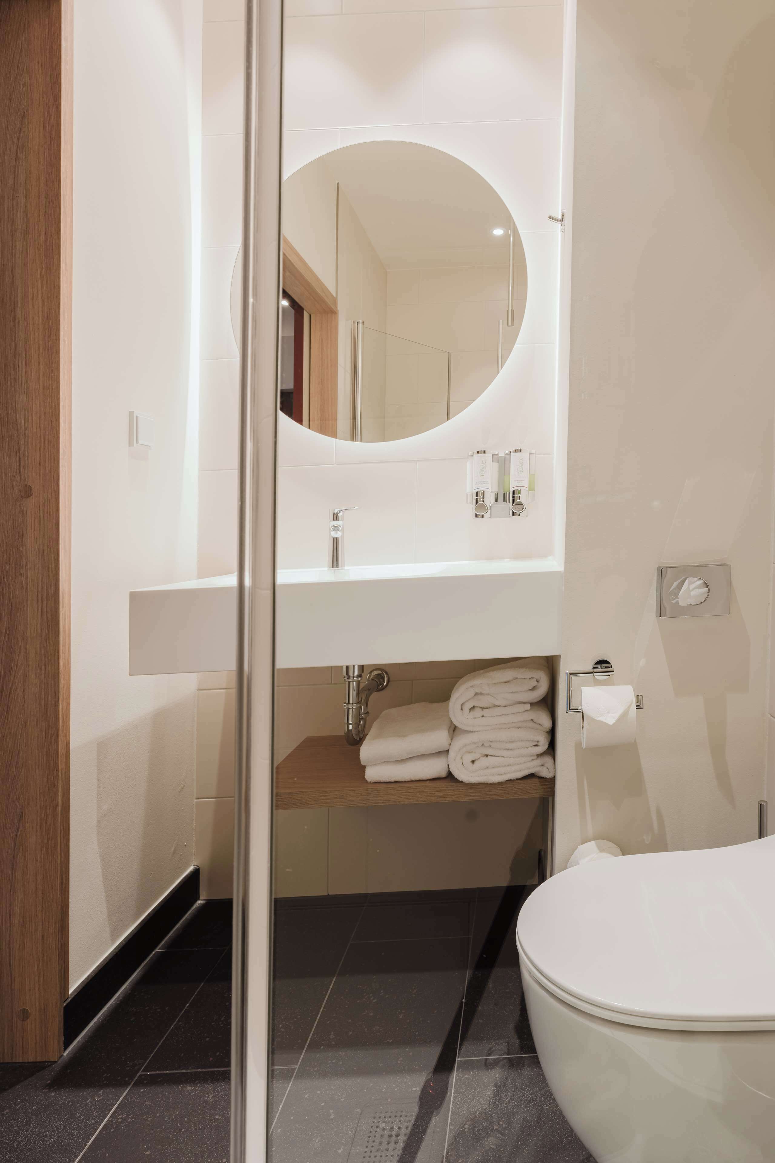 Een moderne badkamer met een douchecabine van glas, een ronde spiegel, een witte wastafel met toiletartikelen en een toilet.