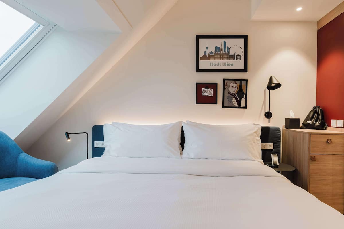 Une chambre d’hôtel avec un lit, un bureau, une chaise, une lampe, une commode et des tableaux encadrés au mur.