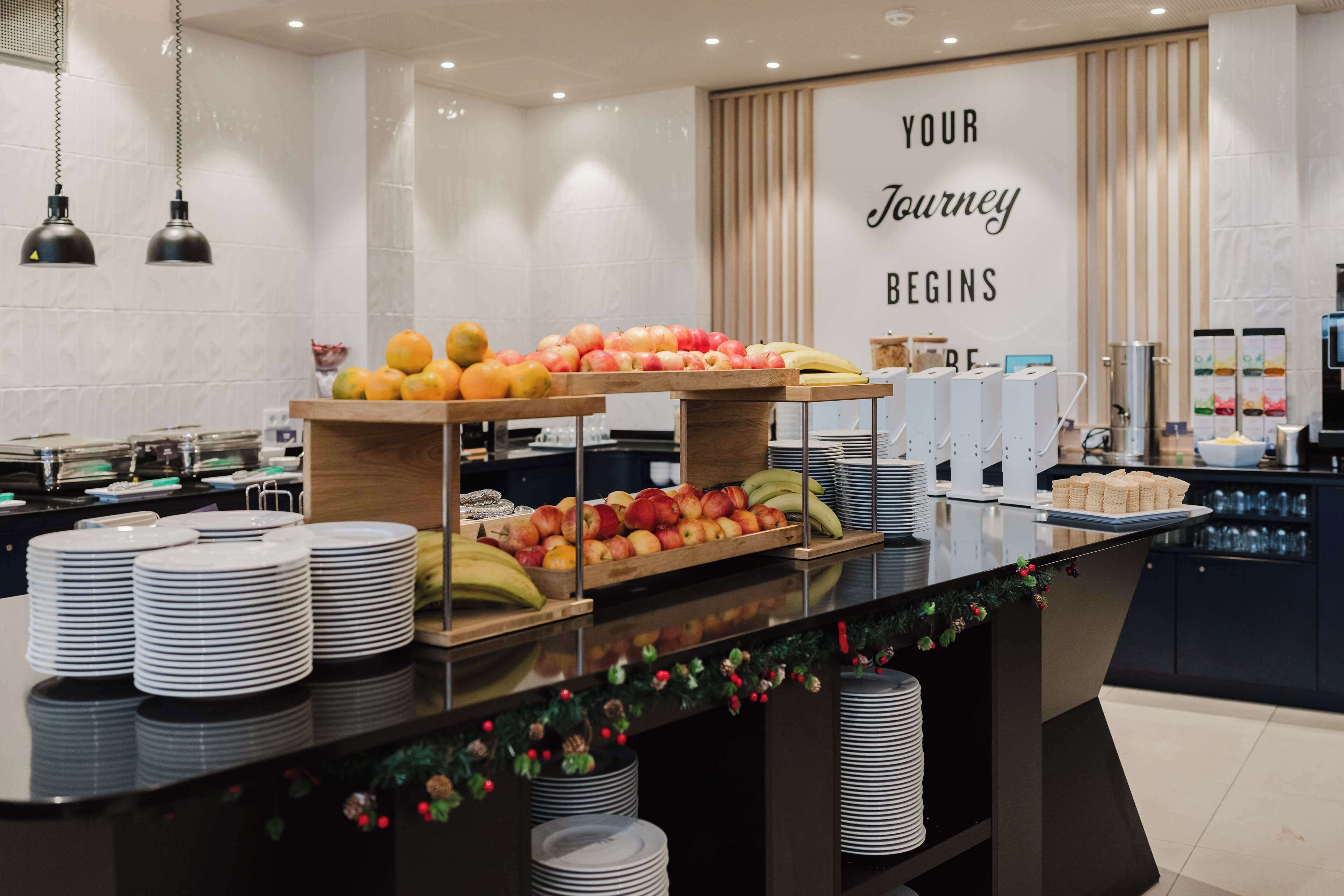 Montaje de bufé con diversas frutas y platos apilados, un cartel que dice "YOUR Journey Begins", y un área de cocina moderna al fondo.