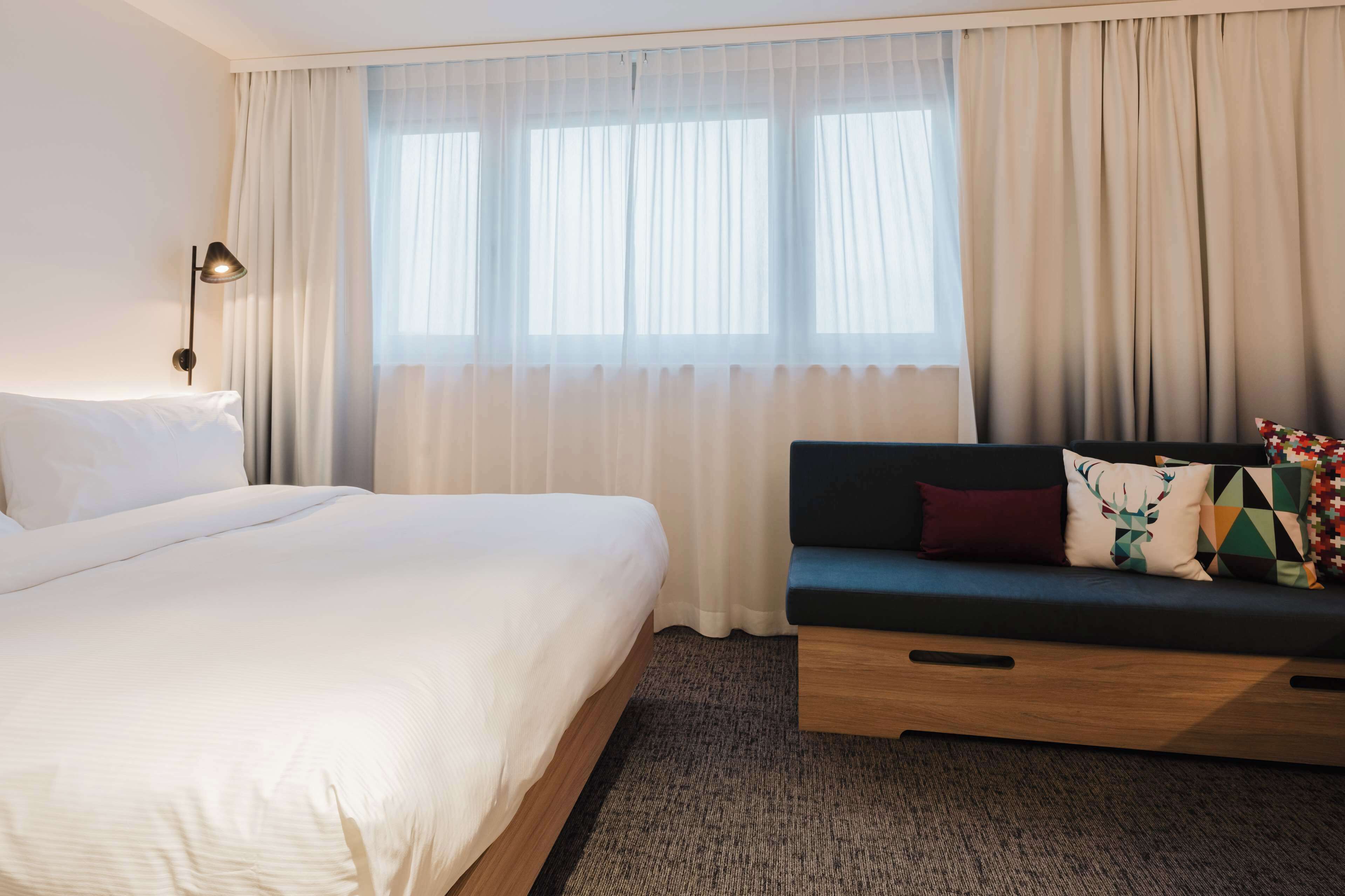 A hotel room with a large bed, a sofa, and a window with sheer curtains.