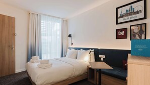 Et hotelværelse med en seng, en sofa, et skrivebord og et bybillede på væggen.