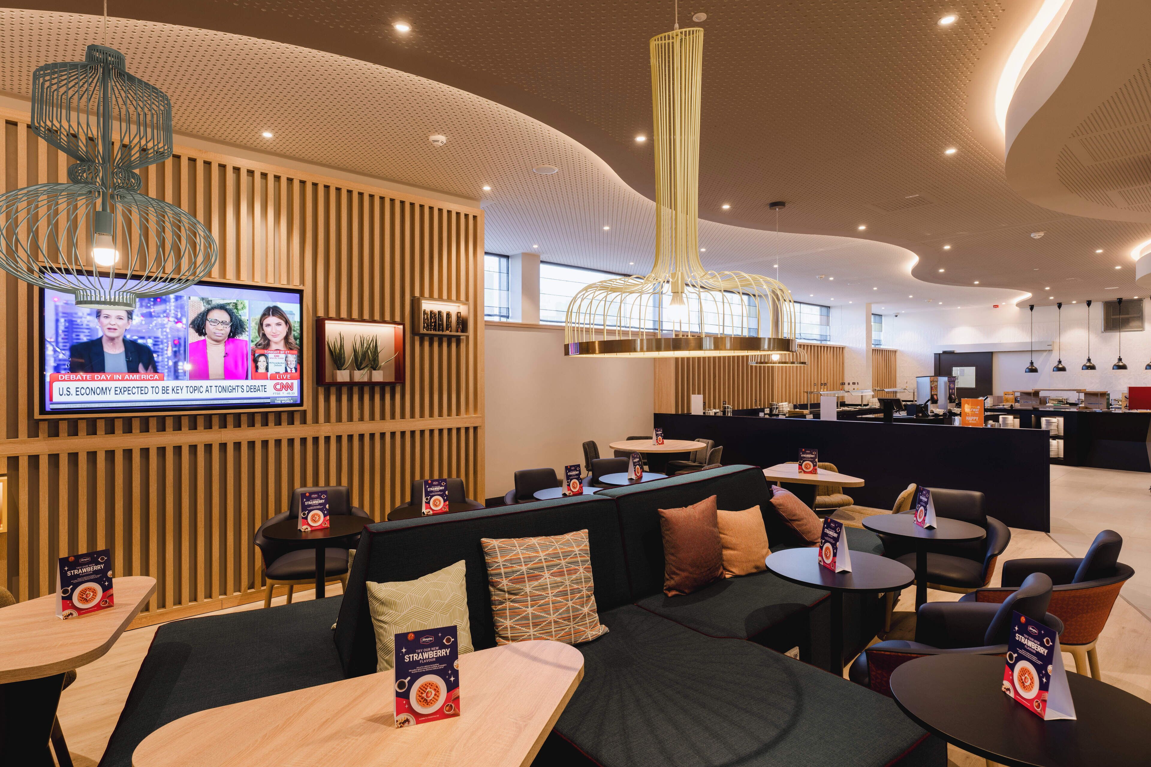 Um hall de hotel moderno, com áreas de estar, uma televisão exibindo um noticiário e um design de teto contemporâneo.