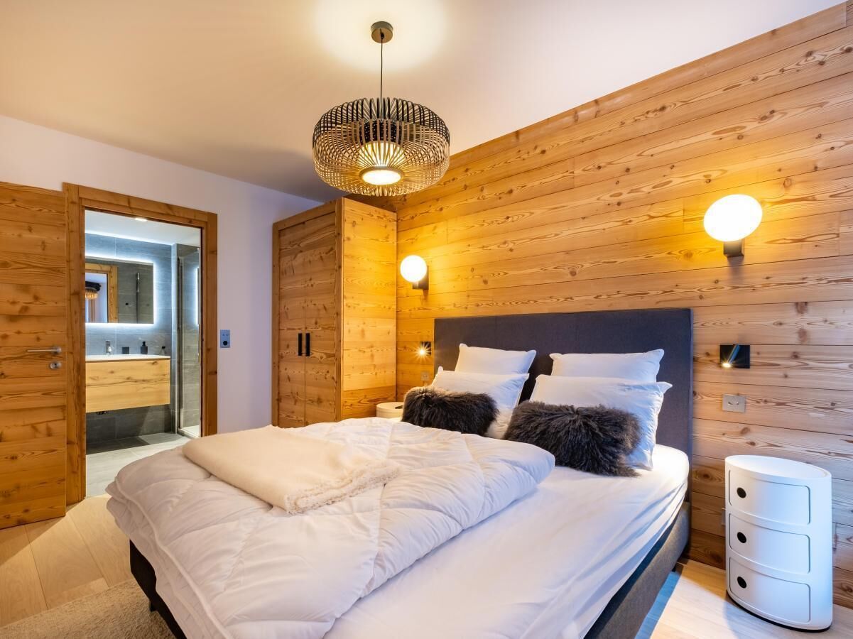Un dormitorio moderno con cabecera de madera, una cama grande con ropa blanca de cama, dos luces de lectura fijadas a la pared y una mesita de noche con lámpara.