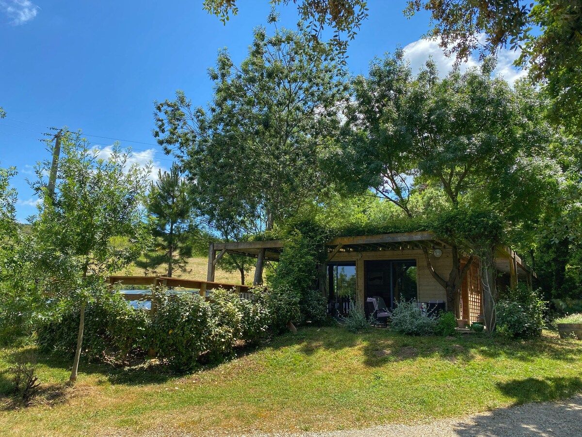 Un chalet rustique doté d’une terrasse en bois, entouré d’une végétation luxuriante et surplombant un ciel bleu azur.
