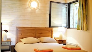A bedroom with a wooden bed, bedside tables, a bench, a window with curtains, and a wall-mounted light fixture. - Ecolodge calme & piscine privée (Roquetaillade-et-Conilhac)