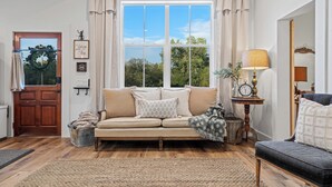 A cozy living room with a beige sofa, a dark armchair, and a wooden coffee table. A large window with curtains allows natural light to fill the space.