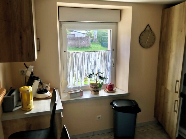 A kitchen with a window, a clock, a trash bin, and a small plant on the windowsill.