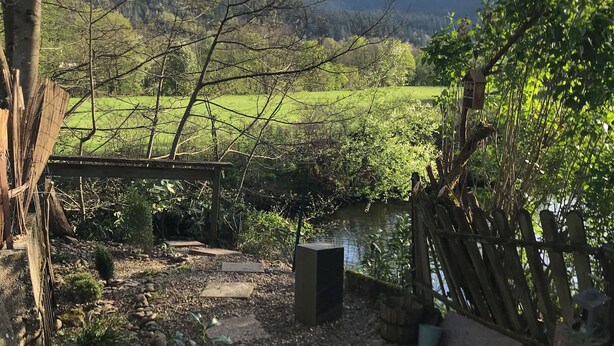 Ein Garten mit Teich, einem Kiesweg, einem Holzzaun und Blick auf ein grünes Feld und Berge in der Ferne.